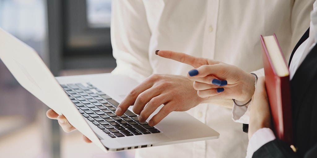 One person is holding a laptop while another person points at the screen, an example of siblings working together.