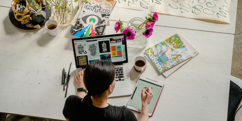 A graphic designer works on their tablet and laptop on a colorfully cluttered desk